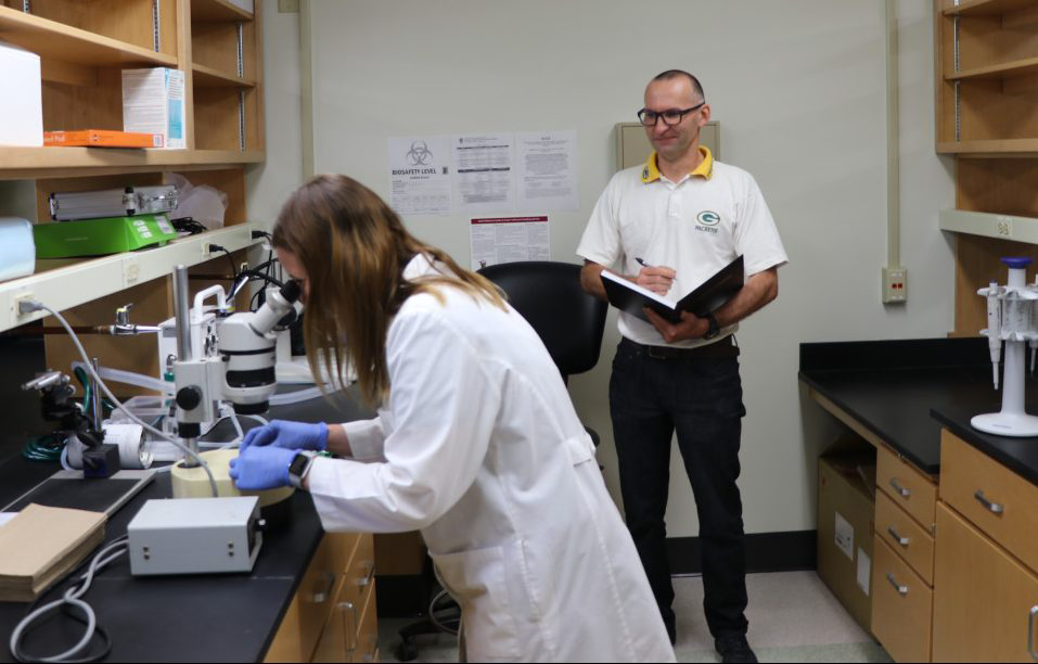A woman looks through a microscope while a man stands behind writing in a notebook